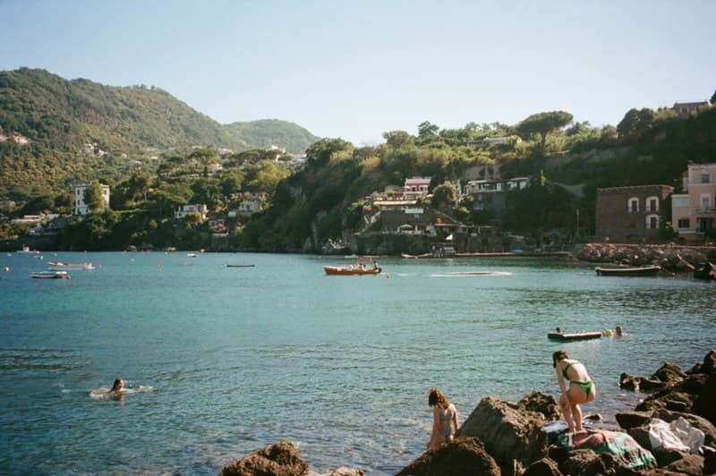 Swimmers in a turquoise cove below a green hillside village